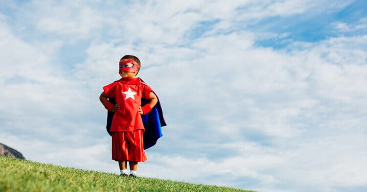 Young boy dressed as super hero, standing on grassy hill, blue sky with clouds behind him. Hands on hips, proud pose.