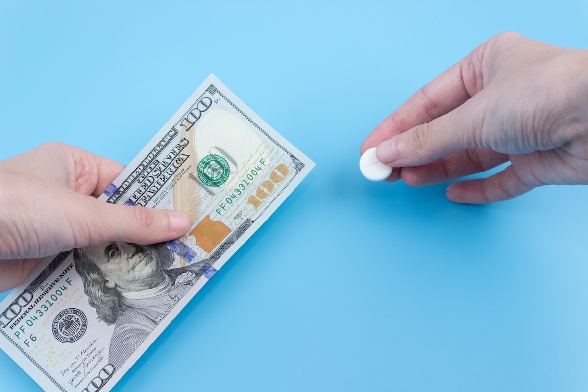Close-up of two hands over a blue surface. One is holding a 100 dollar bill, the other is holding a white pill.